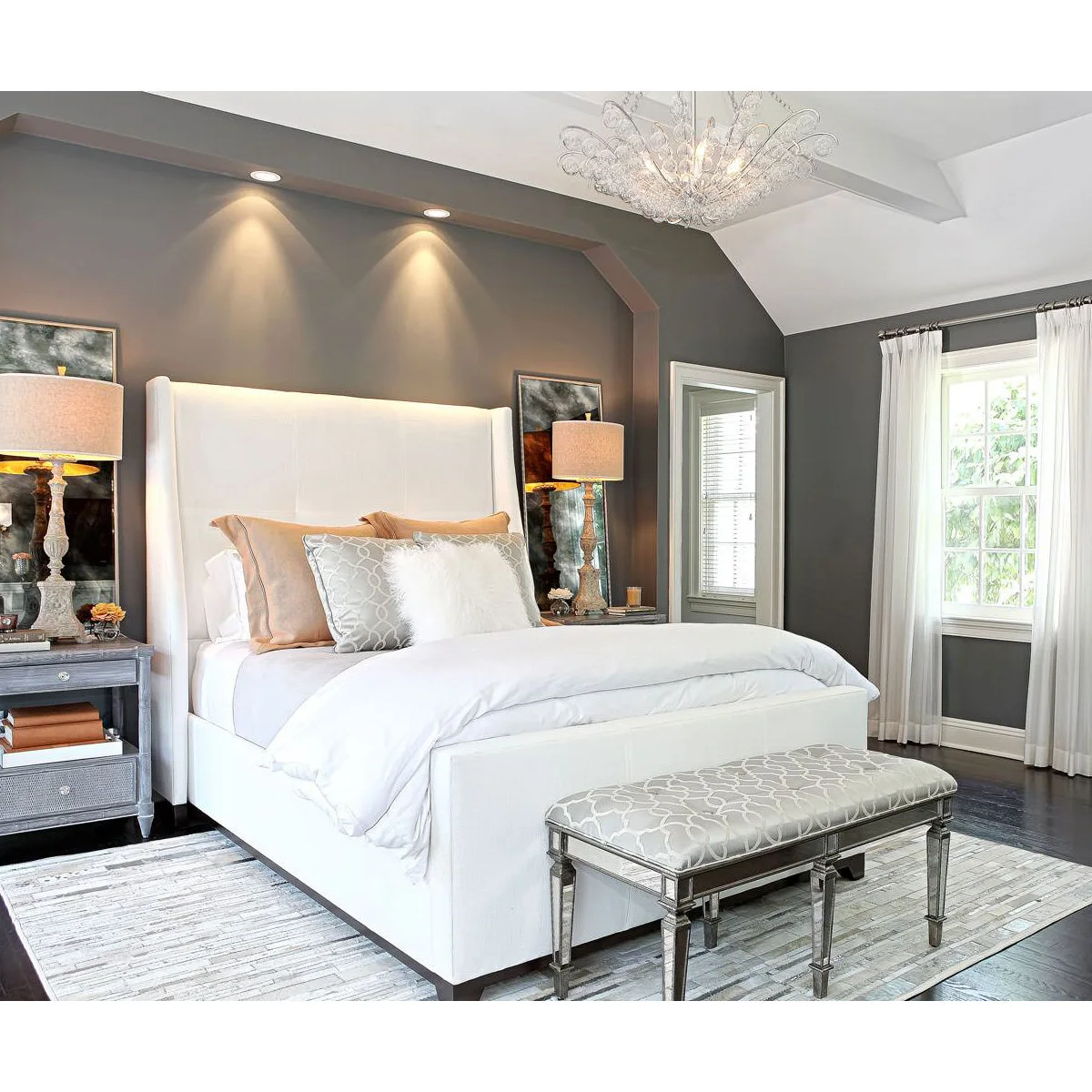 Mid-tone grey bedroom wall behind a tall white bed, paired lamps and patterned bench