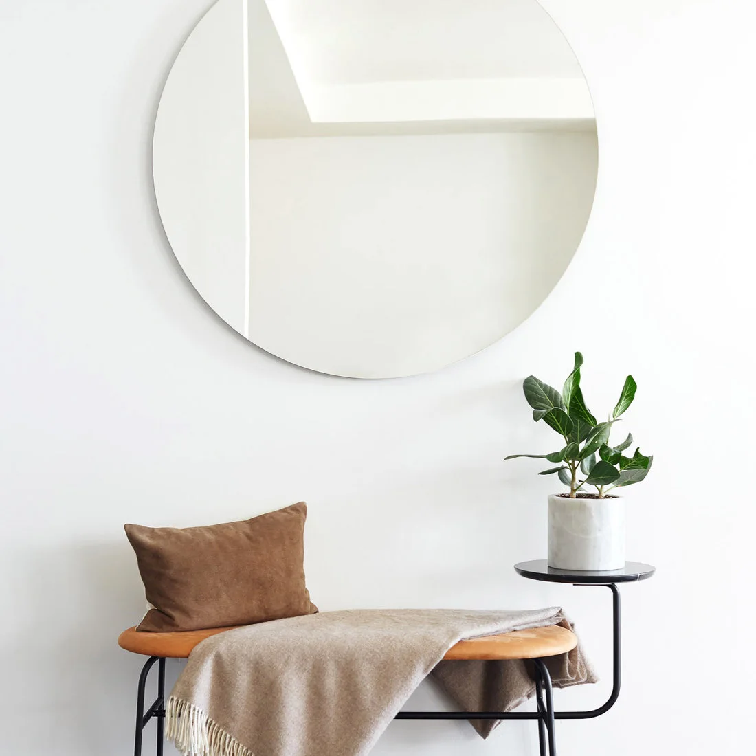 A slim tan bench with black legs and an attached round side surface sits below a round mirror with a pale throw and plant.
