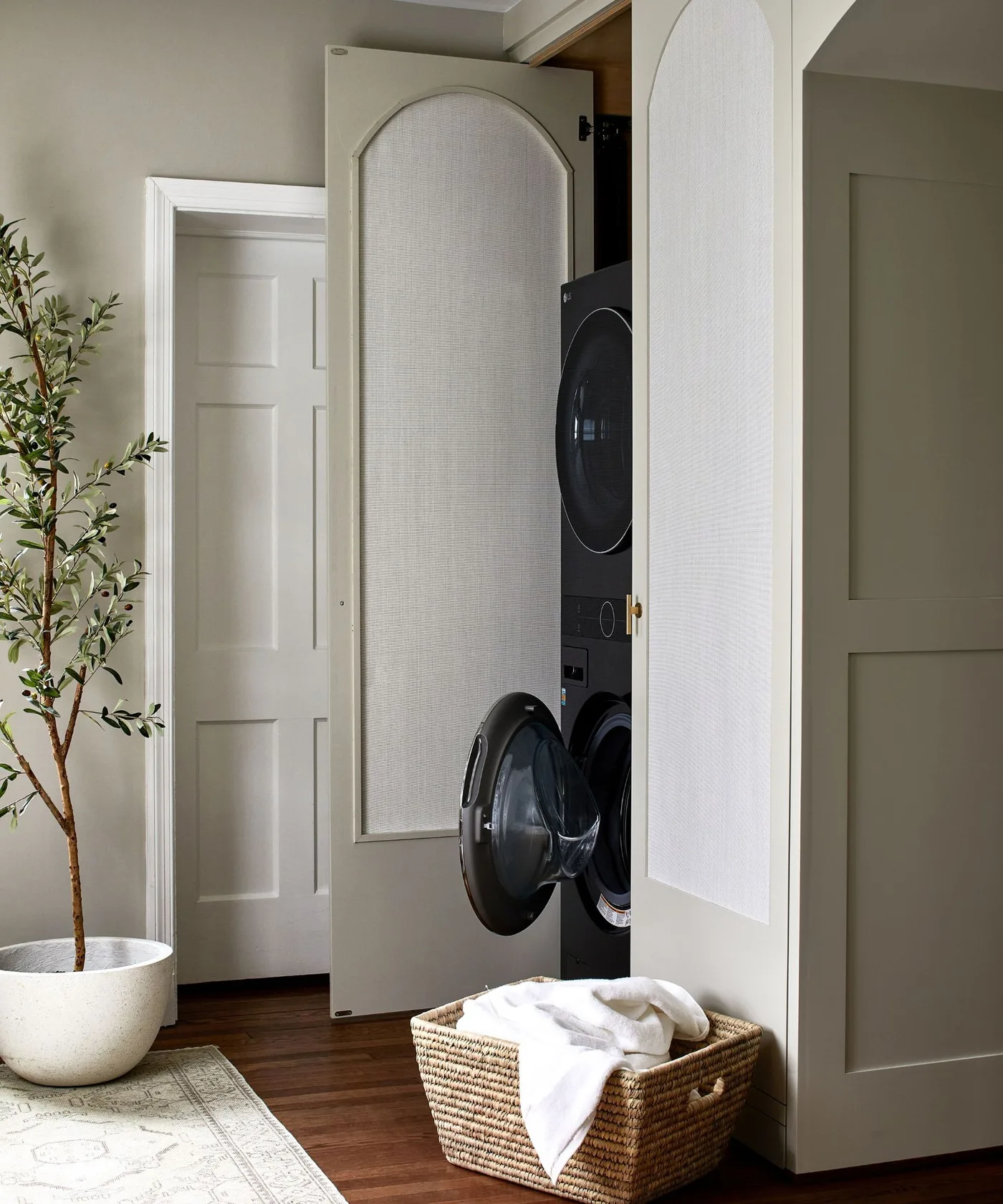 Pale cabinet doors with curved inset panels open around stacked dark laundry machines, with a woven basket on the floor