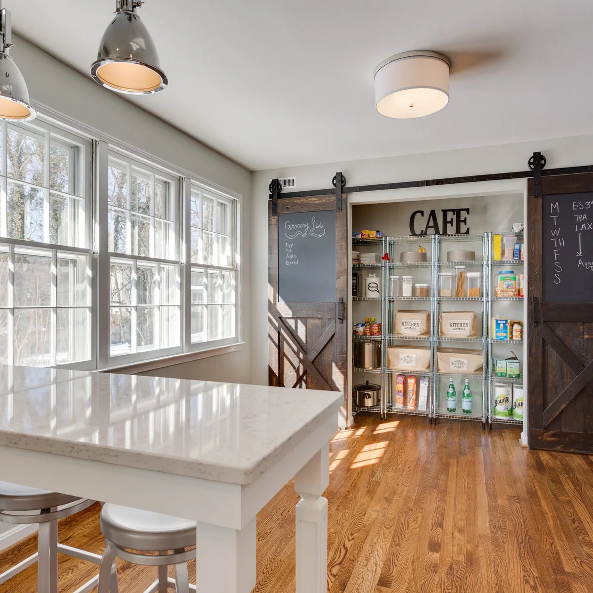 Dark sliding doors with chalkboard panels frame a wide pantry recess containing metal shelves, jars, baskets and boxed food.
