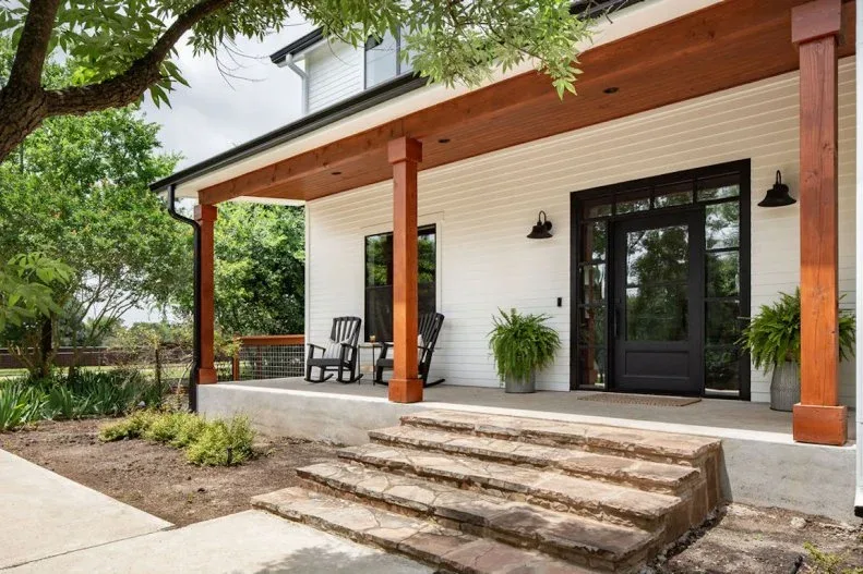 Modern white farmhouse porch with wood posts and ceiling, dark glazed door, black rockers and broad steps