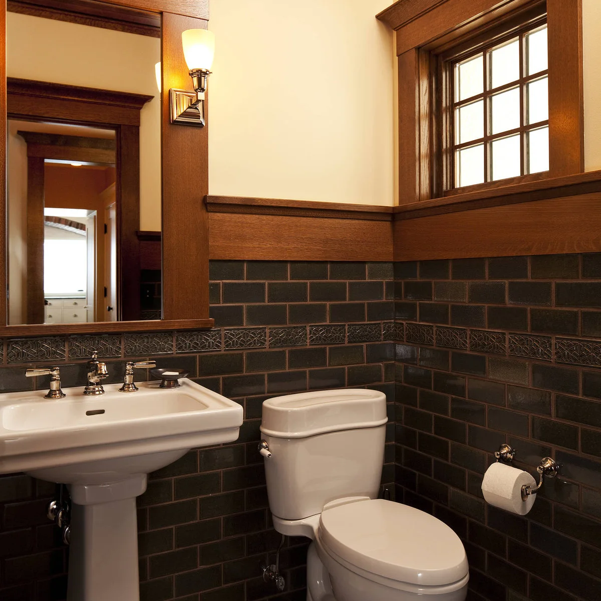 A white pedestal sink sits beside a toilet against dark rectangular tiles with a decorative band and broad wood trim.