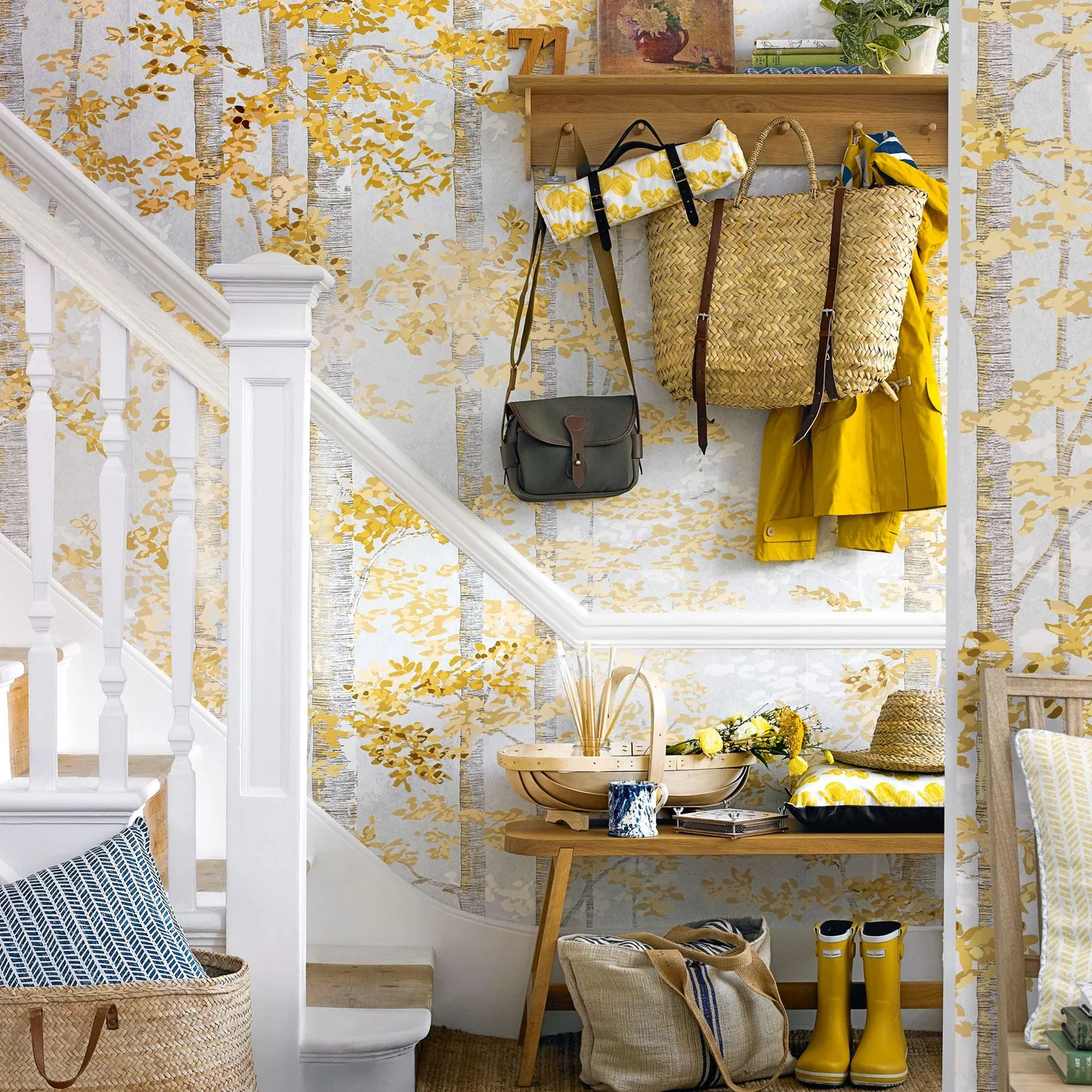 Hallway beside a white staircase with yellow leafy wallpaper, wooden hook shelf, wooden bench, hanging bags and yellow boots.