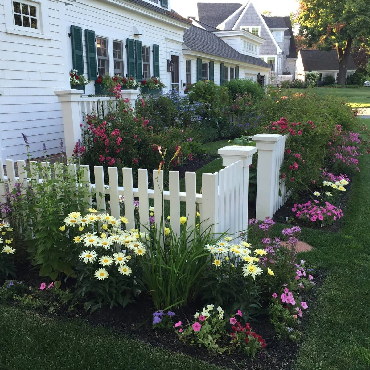 A short white picket fence turns through a flower-filled front garden beside a white house with dark shutters.