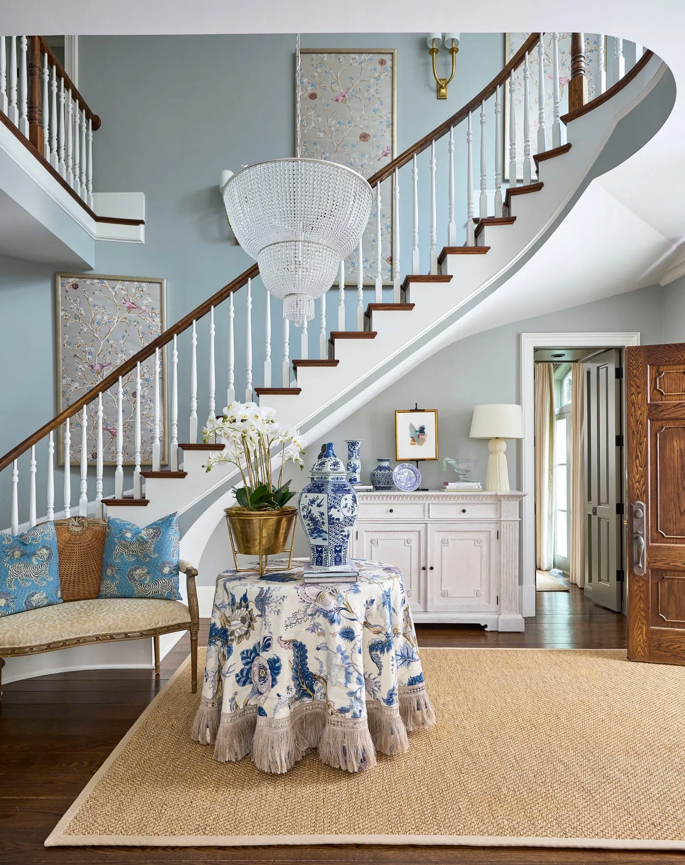Large white tiered beaded chandelier above blue floral-skirted table in pale blue entry with white staircase, woven rug and blue cushions