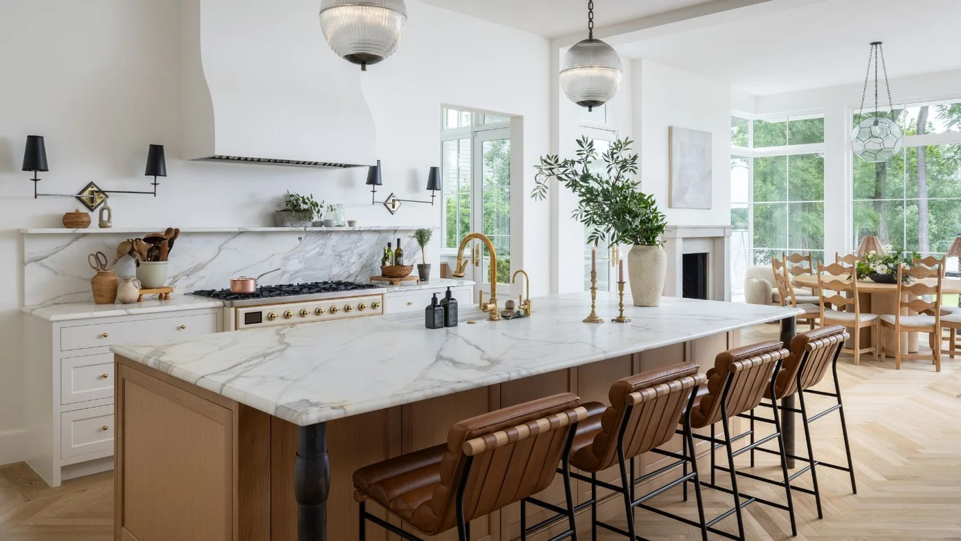 An oak island with a pale veined counter and brown stools faces white cabinets, a white hood and a bright dining area.