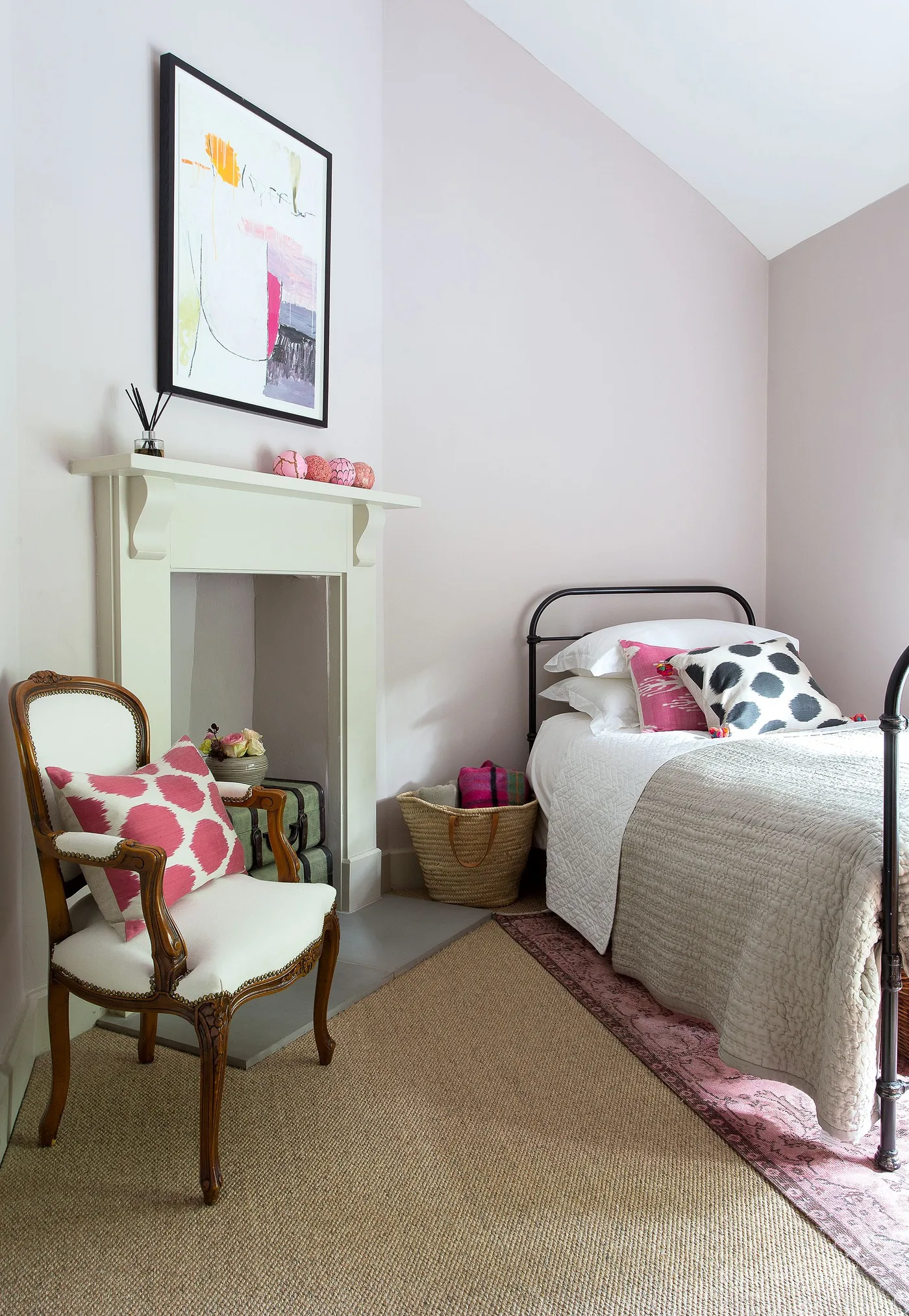 Small pale pink bedroom with a dark metal bed, pale fireplace surround, decorative upholstered chair and bright cushions.