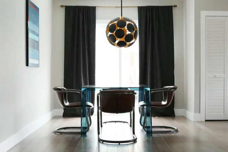 Blue glass dining table with broad transparent sides and dark chairs beneath a black and gold-toned pendant