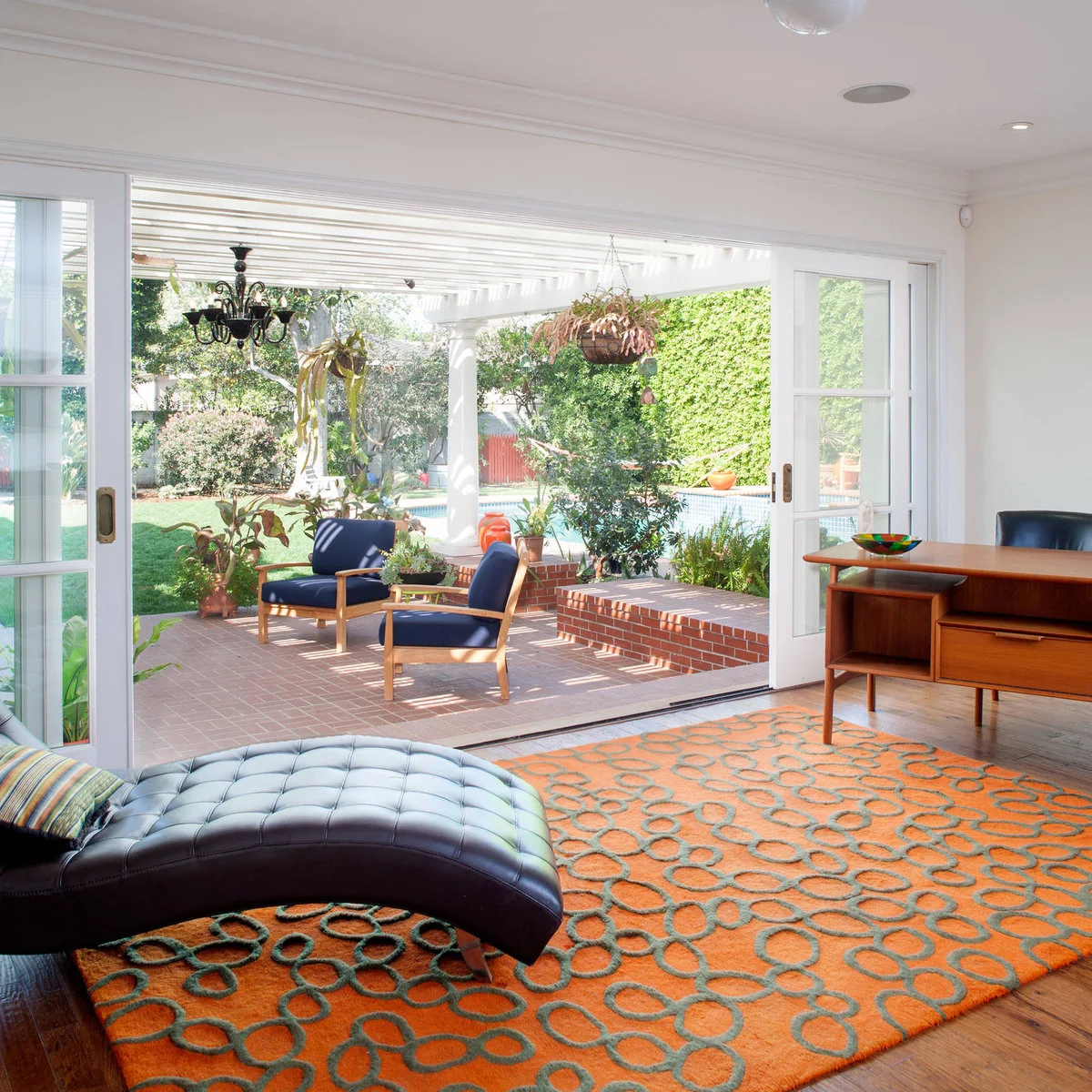 A curved dark chaise and wood desk beside wide sliding doors opening to a garden seating area.