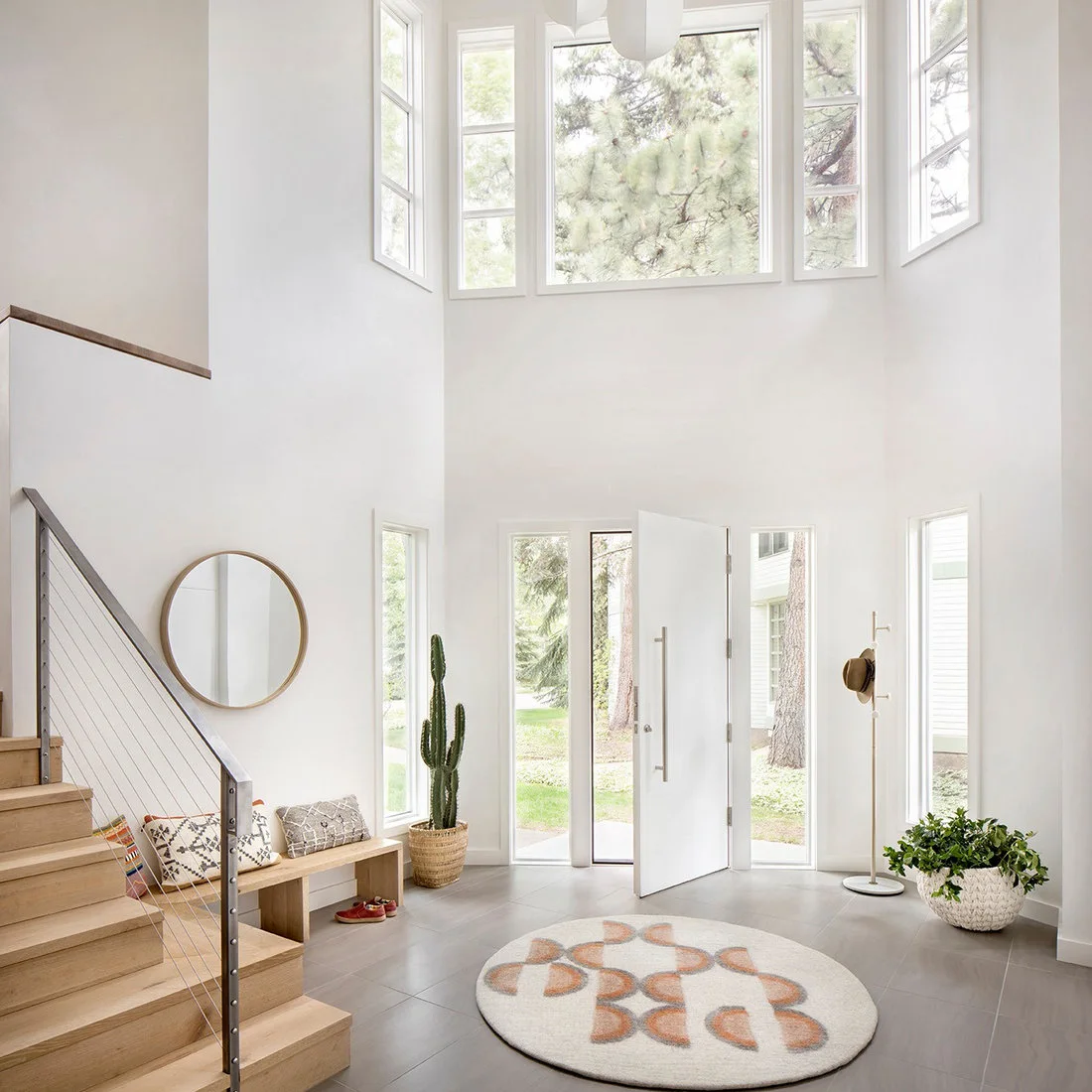 A bright double-height entry has a wood stair, low bench, round mirror and circular patterned rug beside an open white front door.