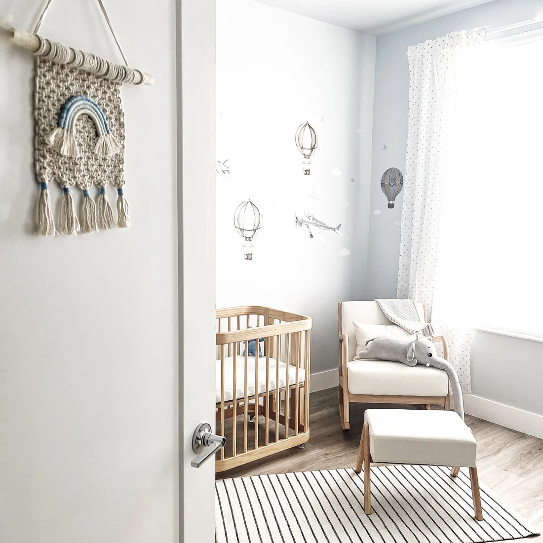 A rounded wood crib and pale upholstered chair with a footstool sit beside a bright window, striped rug and balloon wall illustrations.