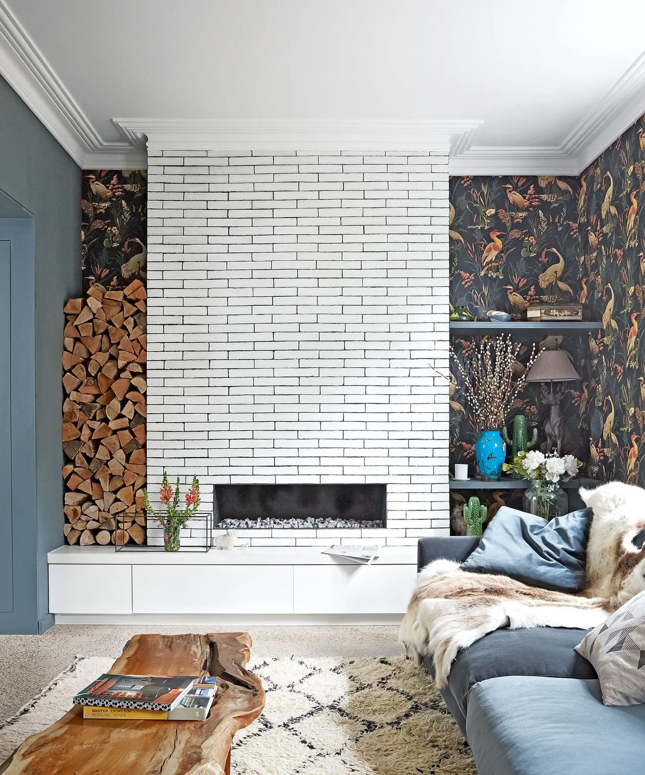 A pale brick fireplace stands beside darker patterned wallpaper and soft seating.