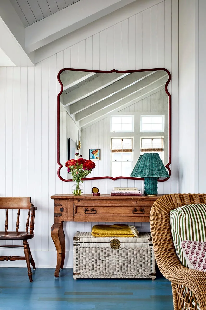 Wood console with green lamp beneath a red-framed mirror, against white paneling and blue floorboards with woven chest below