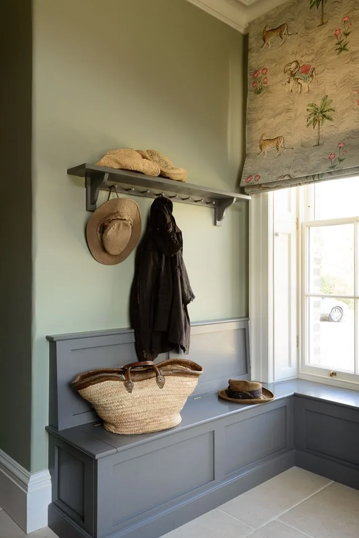 Gray paneled mudroom bench below a peg shelf beside green walls and a patterned window blind.