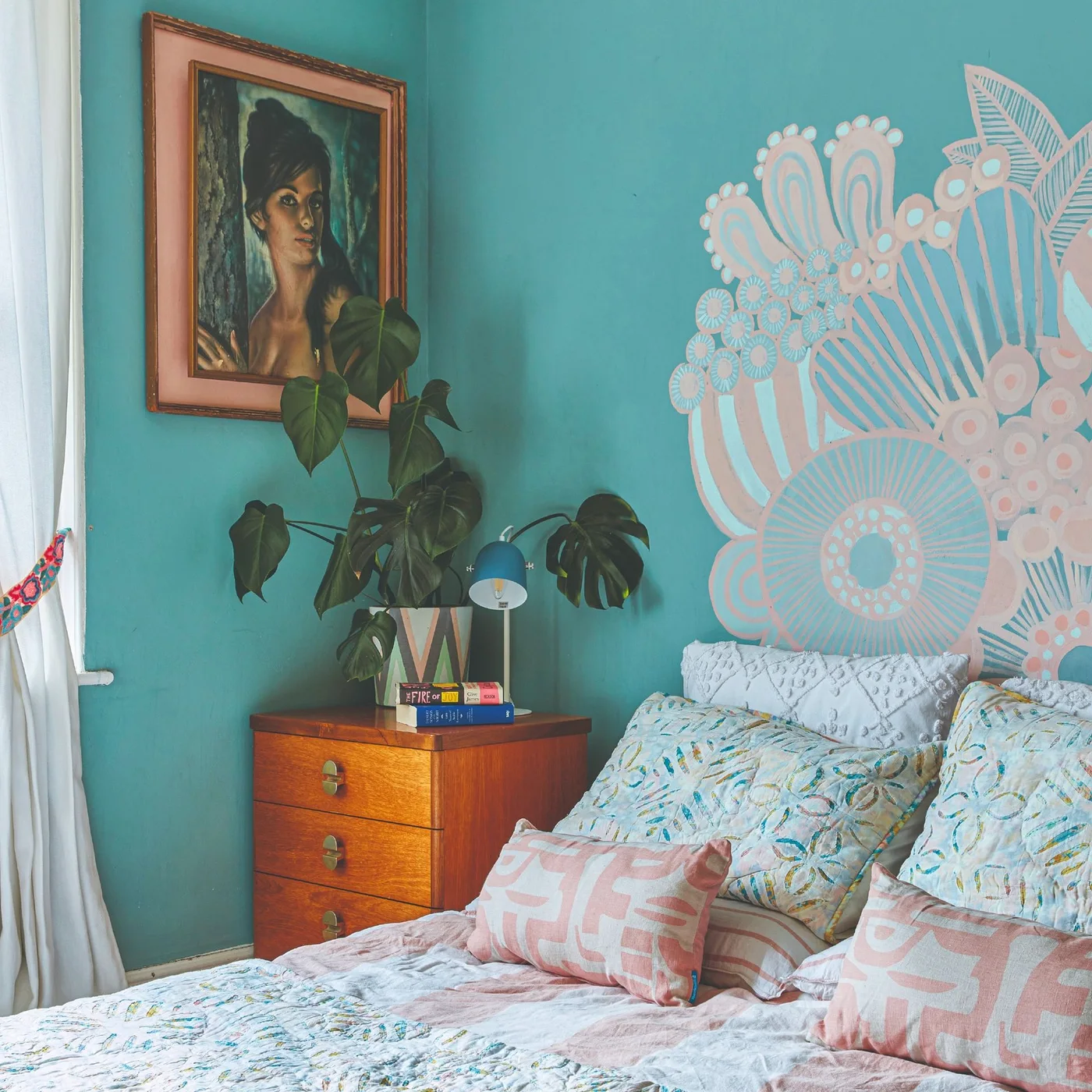 A pale decorative mural spreads above a bed against turquoise walls beside a wood drawer chest, a leafy plant and a framed portrait.