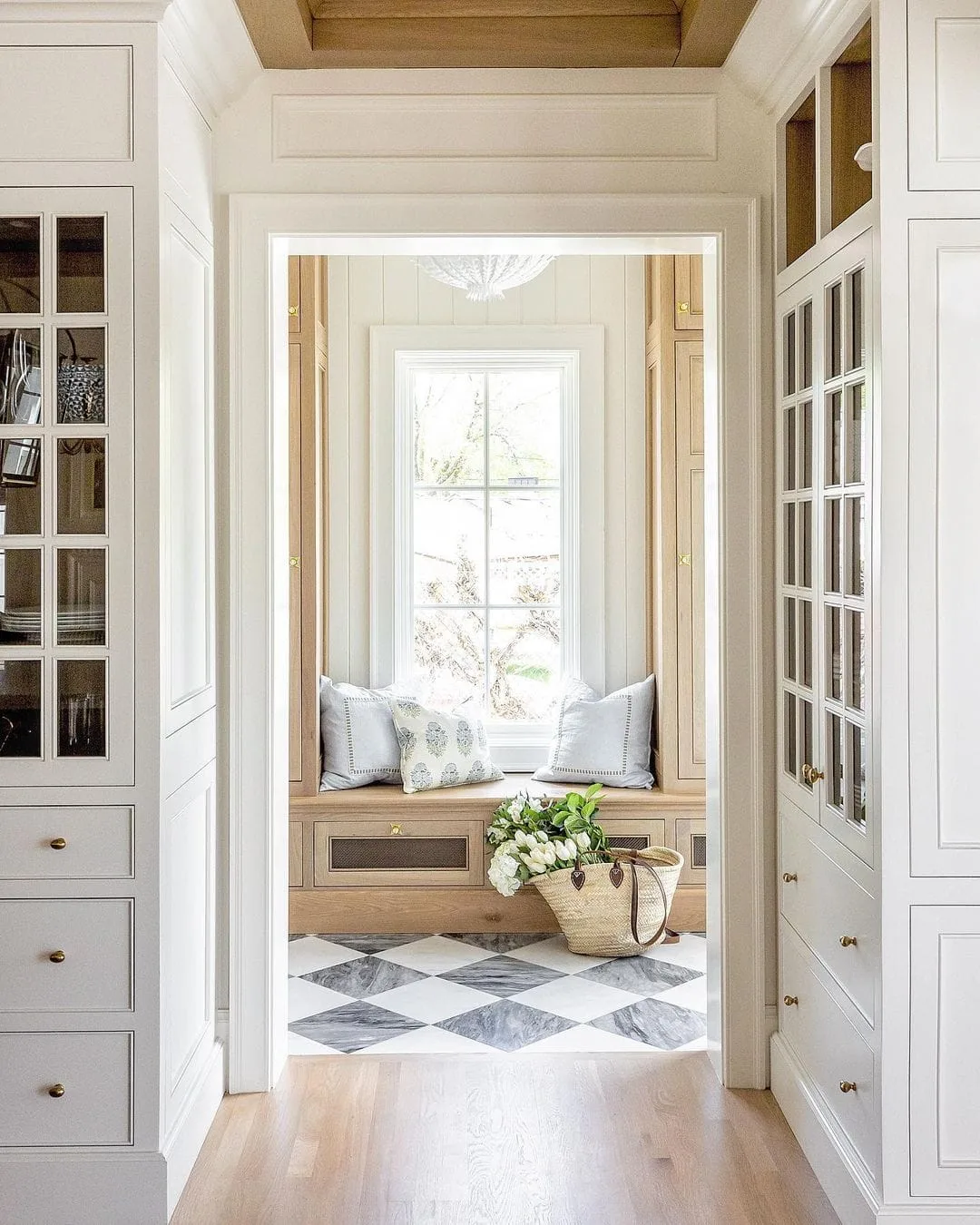 Window seat between glass-front mudroom cupboards with a pale checkerboard floor and wooden ceiling detail.