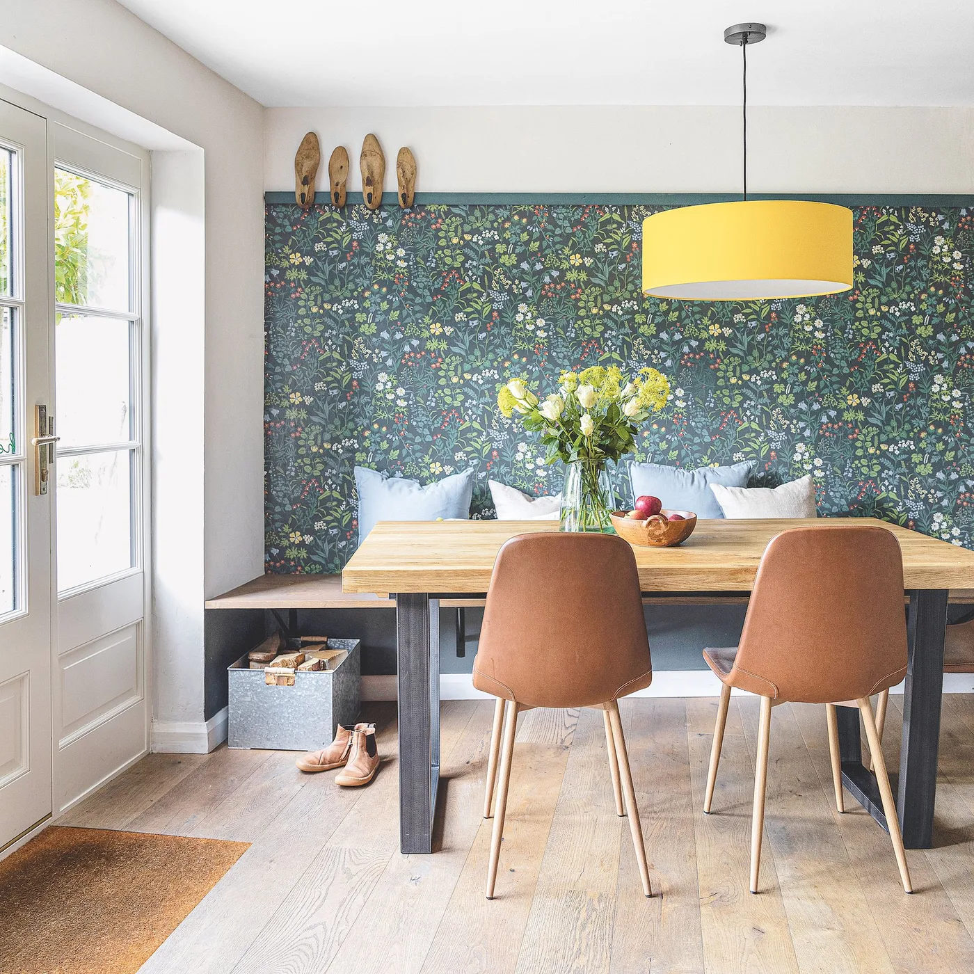 Green botanical wallpaper sits behind a built-in bench and wooden table beneath a yellow pendant beside warm-toned chairs
