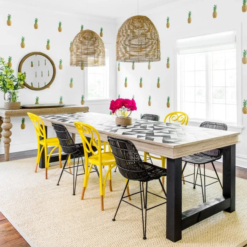 Dining room with pineapple wallpaper, yellow and black chairs, patterned tabletop detail, two woven pendants and a round wall mirror