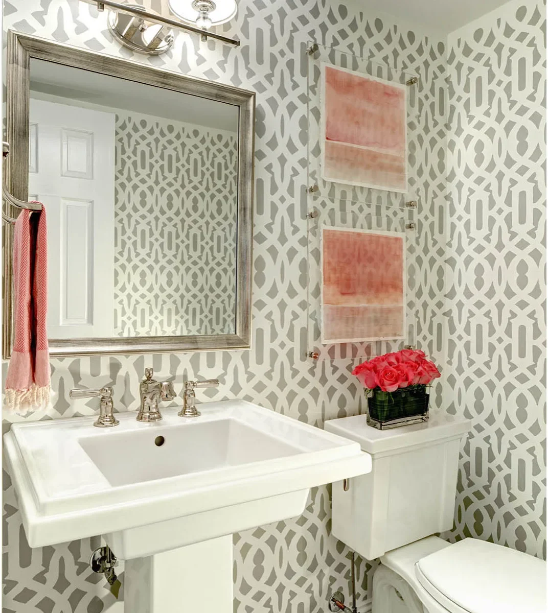 A white pedestal sink sits against gray and white patterned walls, with coral towels, two coral-toned pictures and a broad framed mirror.