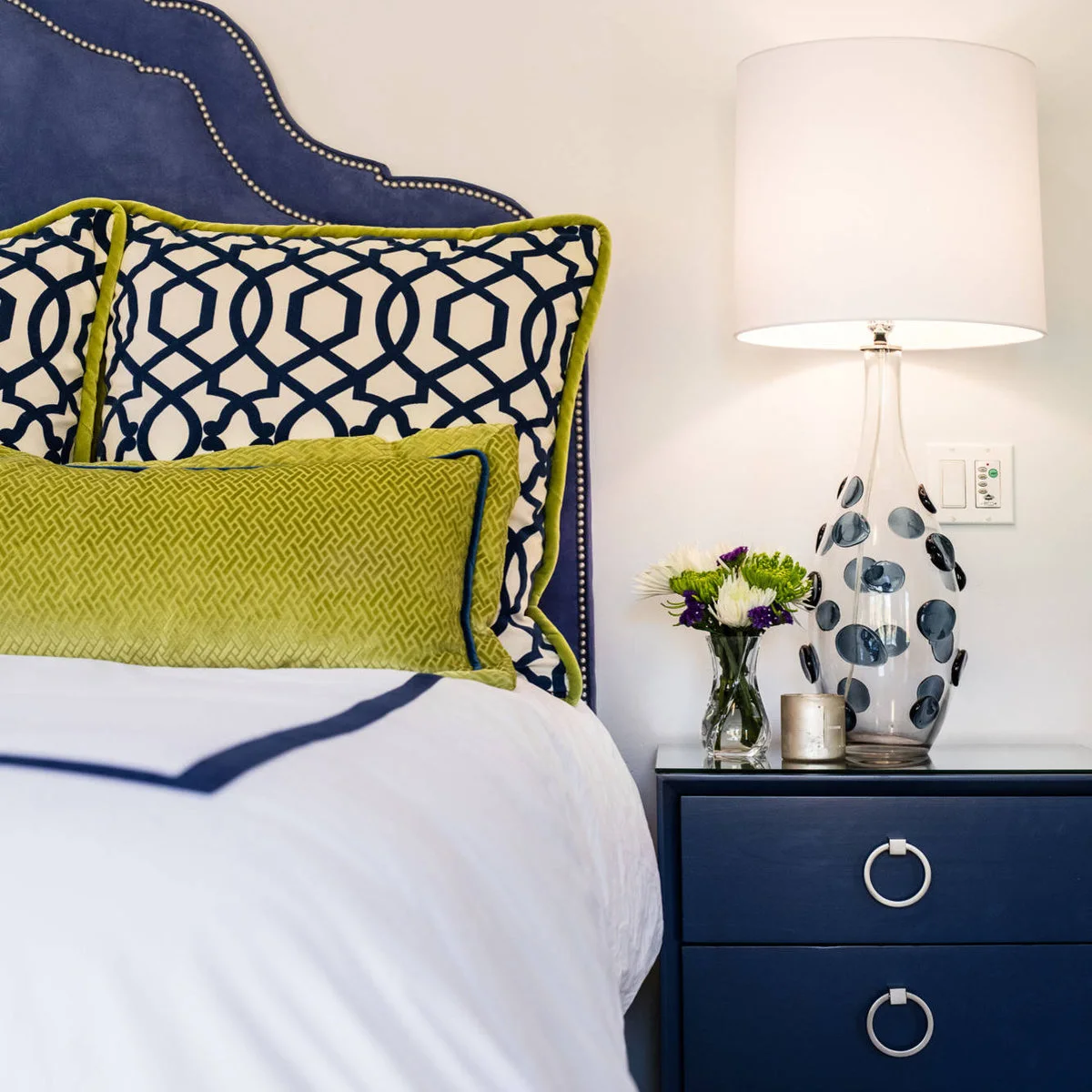 A blue bedside drawer chest with a spotted glass lamp, small flower vase and green-edged patterned pillows.