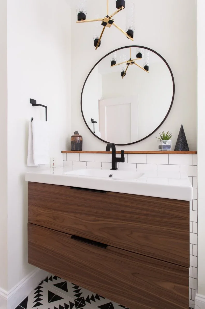 A floating wood drawer vanity with a white basin sits beneath a black round mirror beside black taps, white rectangular wall tiles and a narrow wood ledge