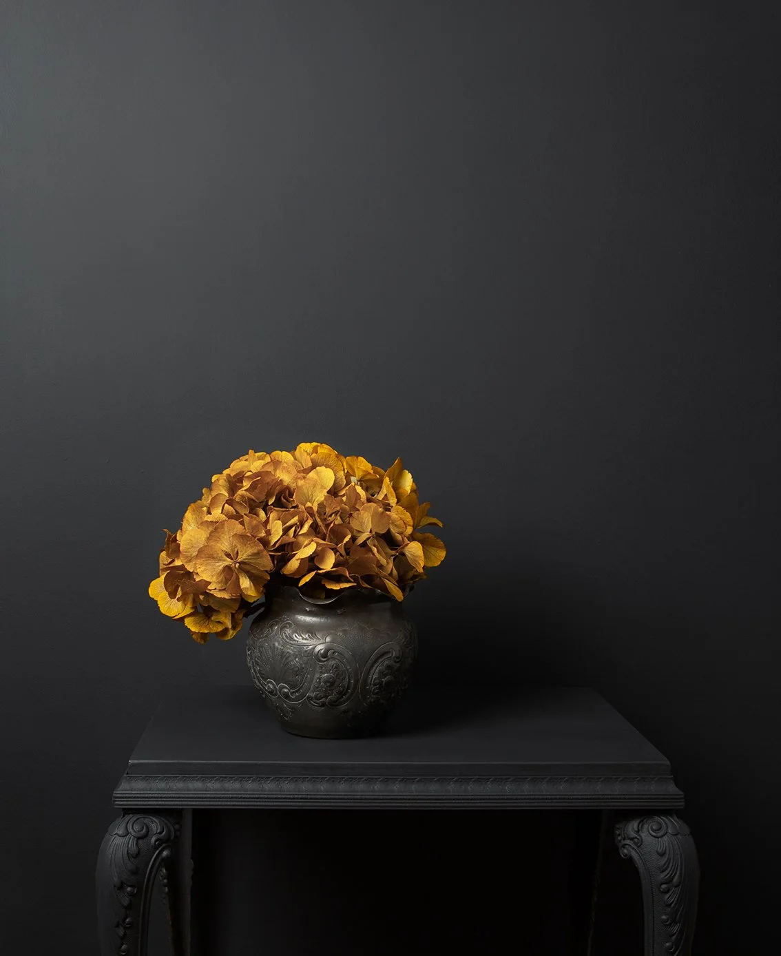 A mustard-colored preserved hydrangea head sits in a small dark patterned pot on a dark table against a charcoal wall.