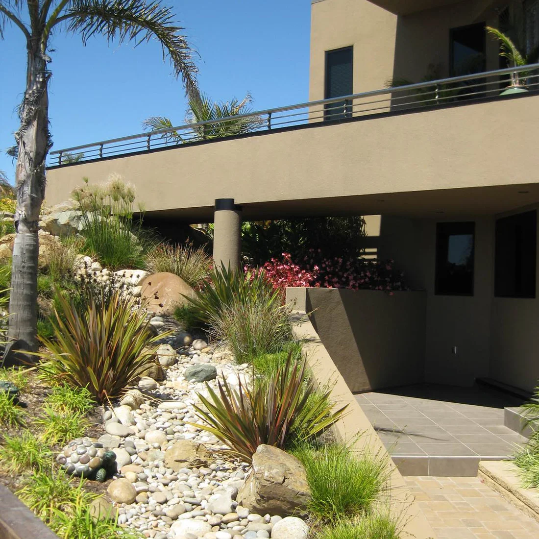 A pale river-rock ribbon curves through spiky planting on a slope beside an exterior staircase