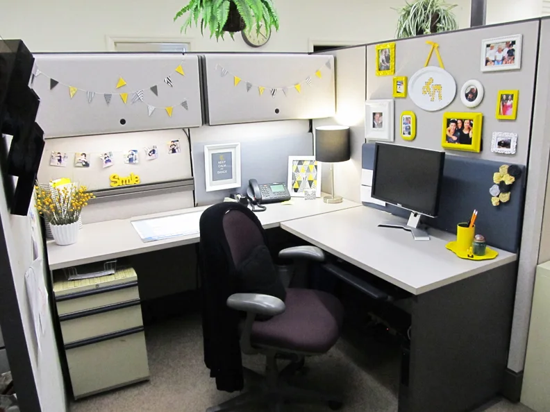 Gray office cubicle with yellow and white picture frames, a short bunting garland, two monitors and a small table lamp.