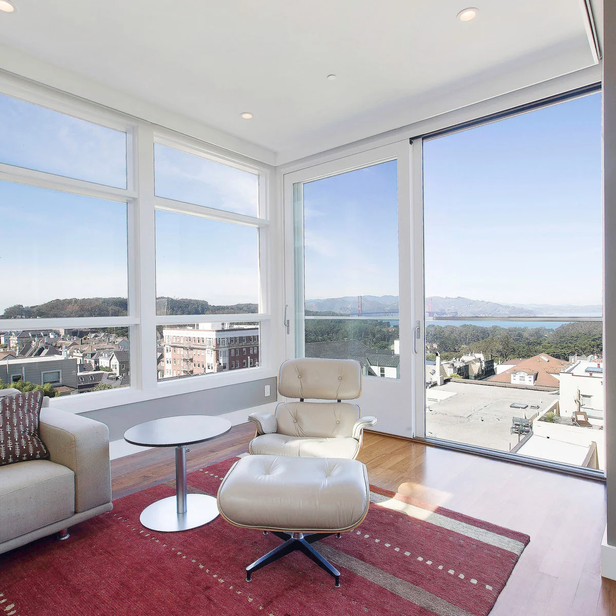Light gray sofa and wall panels surround a cream lounge chair on a burgundy rug beside large windows.