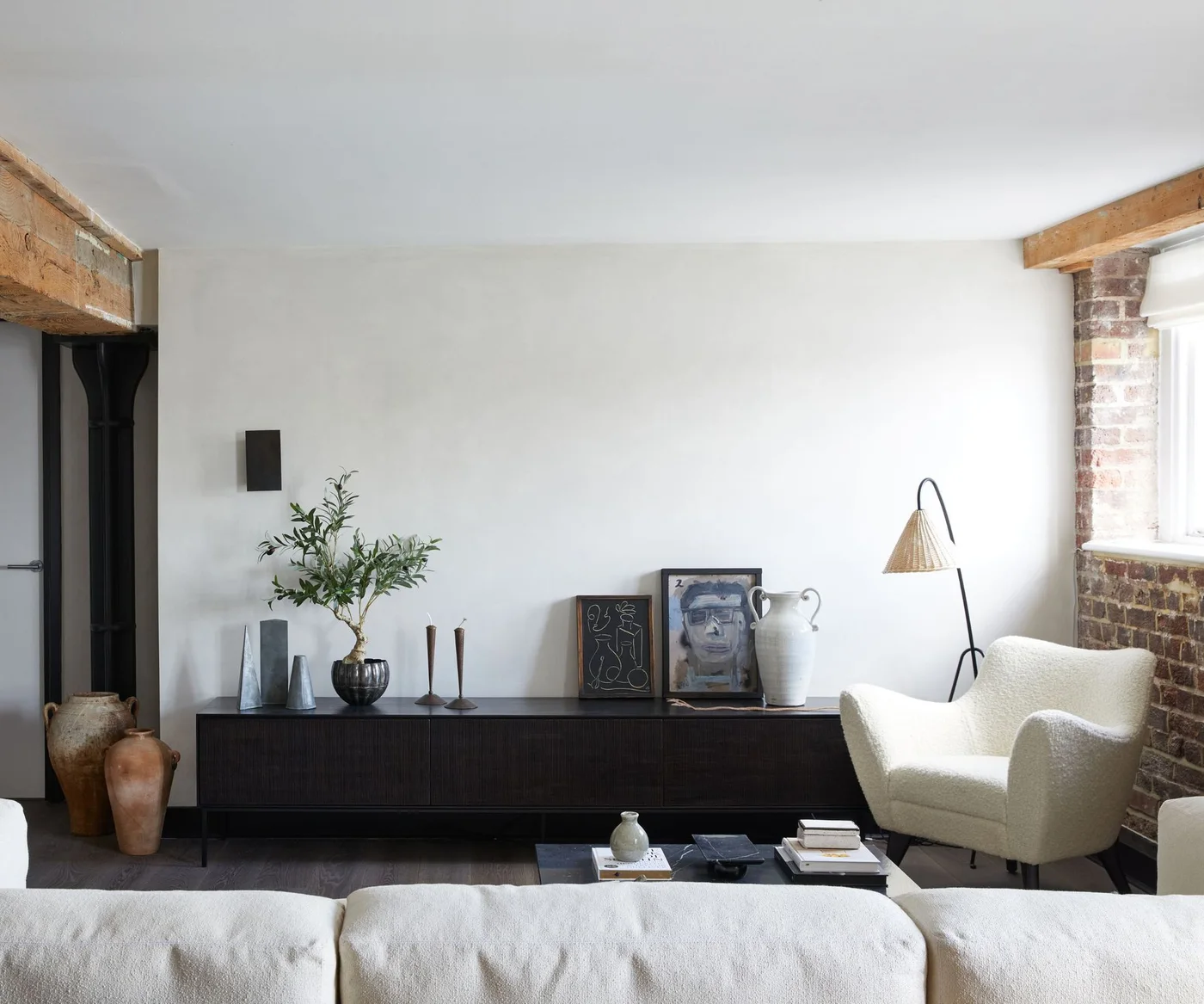 A dark low sideboard stands behind pale textured seating beside exposed brick, a timber beam and a woven-shade floor lamp.
