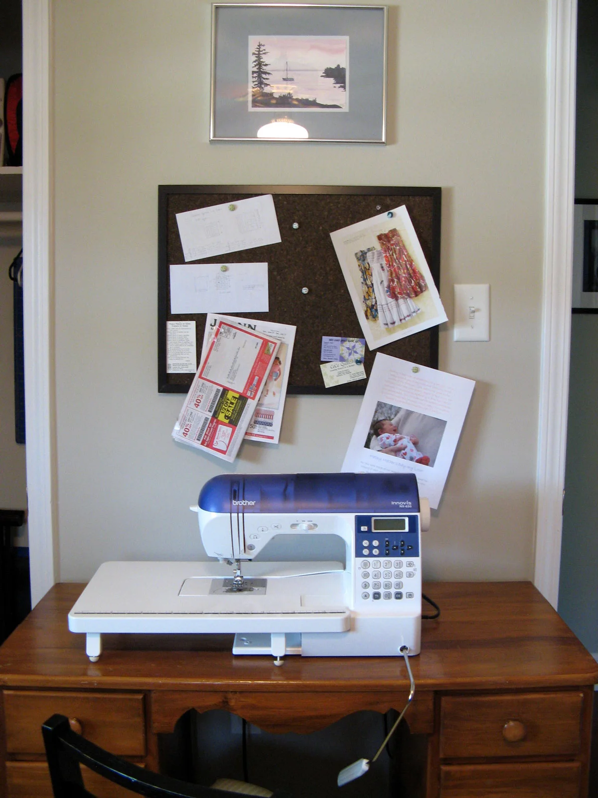 A blue-and-white sewing machine with an extension surface stands on a small wood desk below a framed noticeboard.