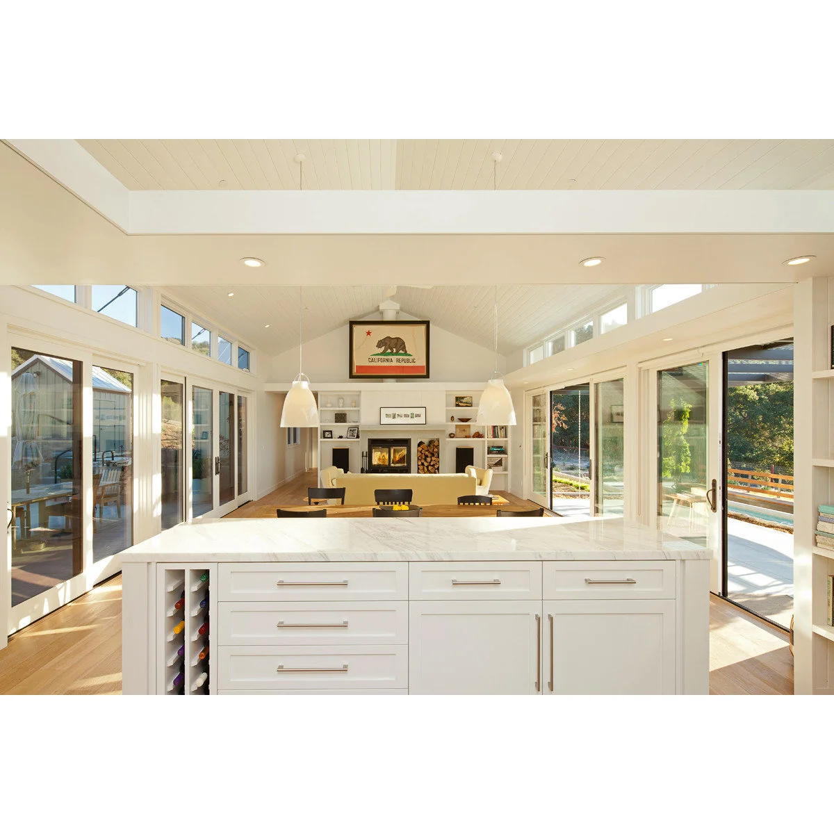 Long pale room seen over a white kitchen island, with a dining table, sofa and fireplace arranged in a line.