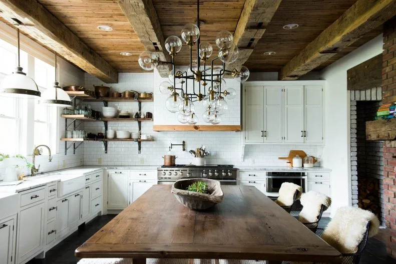 A broad wood table and dark chairs with pale throws stand in a white kitchen beneath heavy timber beams and glass-globe lighting.