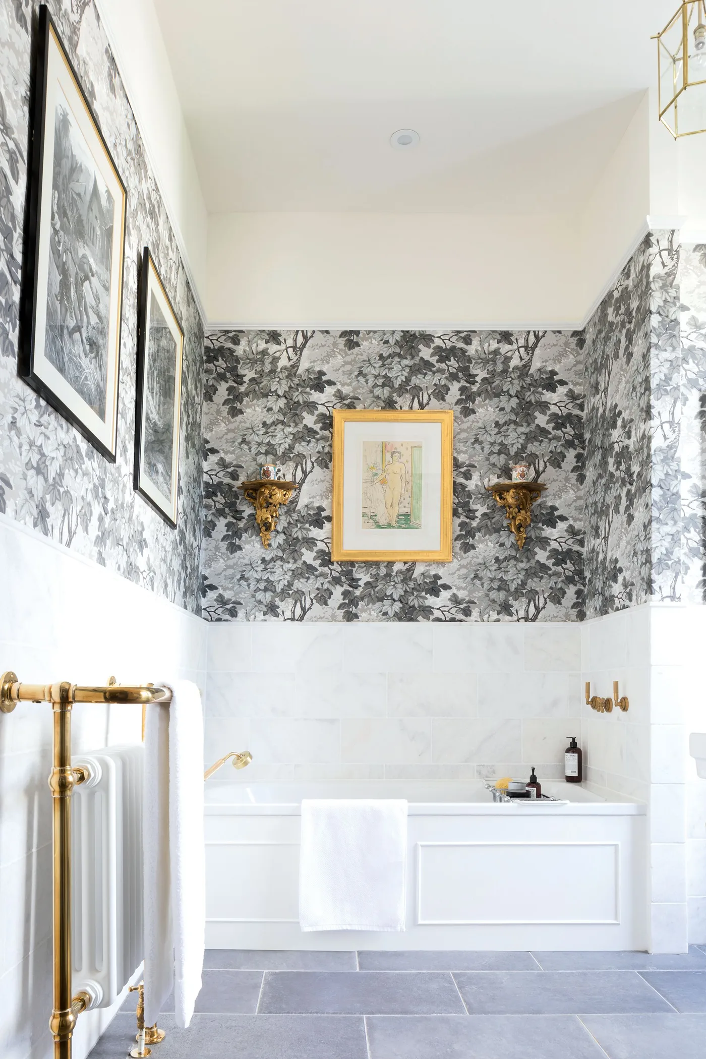 Compact bathroom with a white inset tub, paneled bath front, monochrome wallpaper and warm metal towel rail.