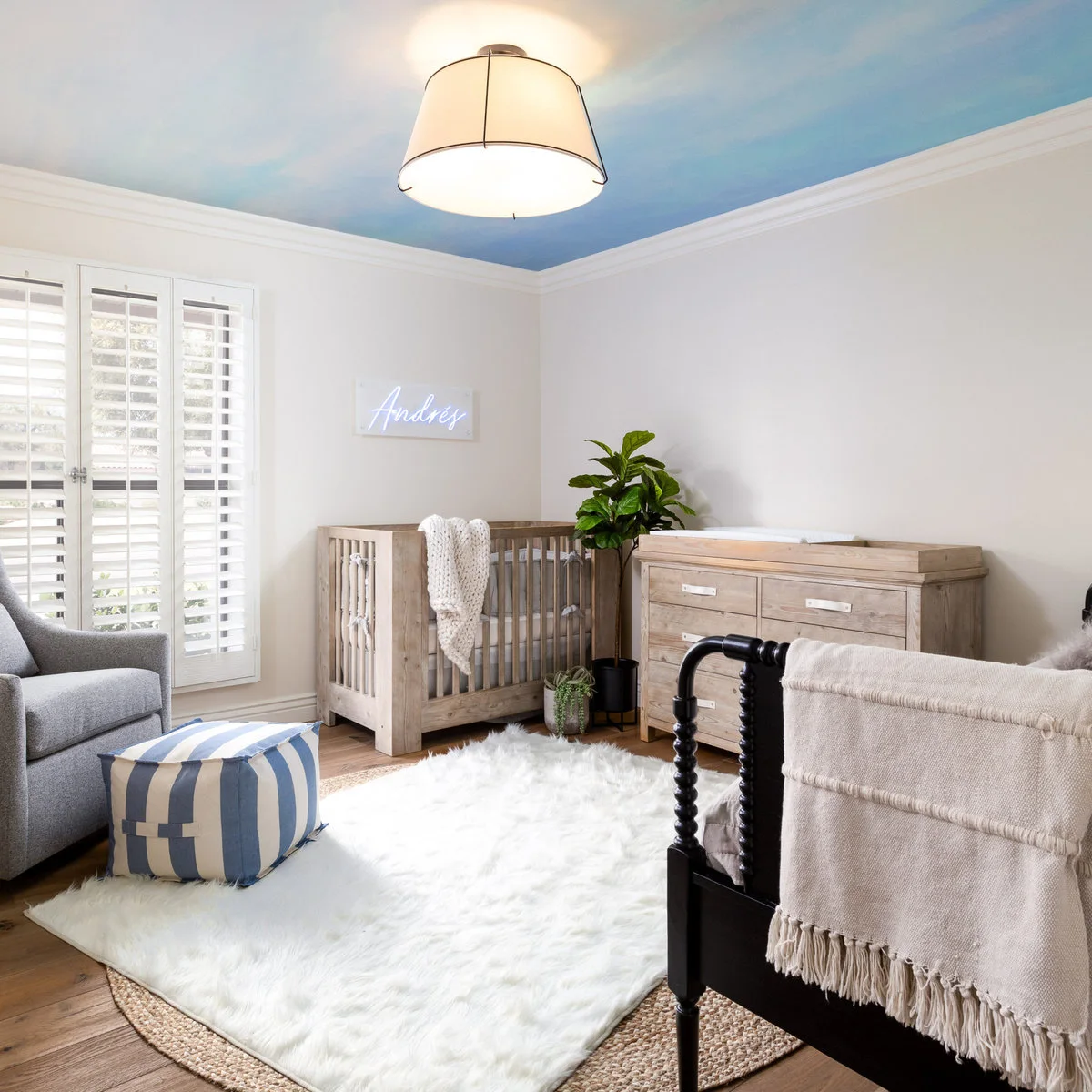Nursery with a blue sky-effect ceiling, wood-colored crib and dresser, gray chair and striped ottoman