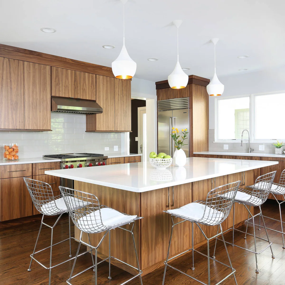 Wire-framed chairs with white seat pads around a pale island in a wood kitchen beneath three white pendants.