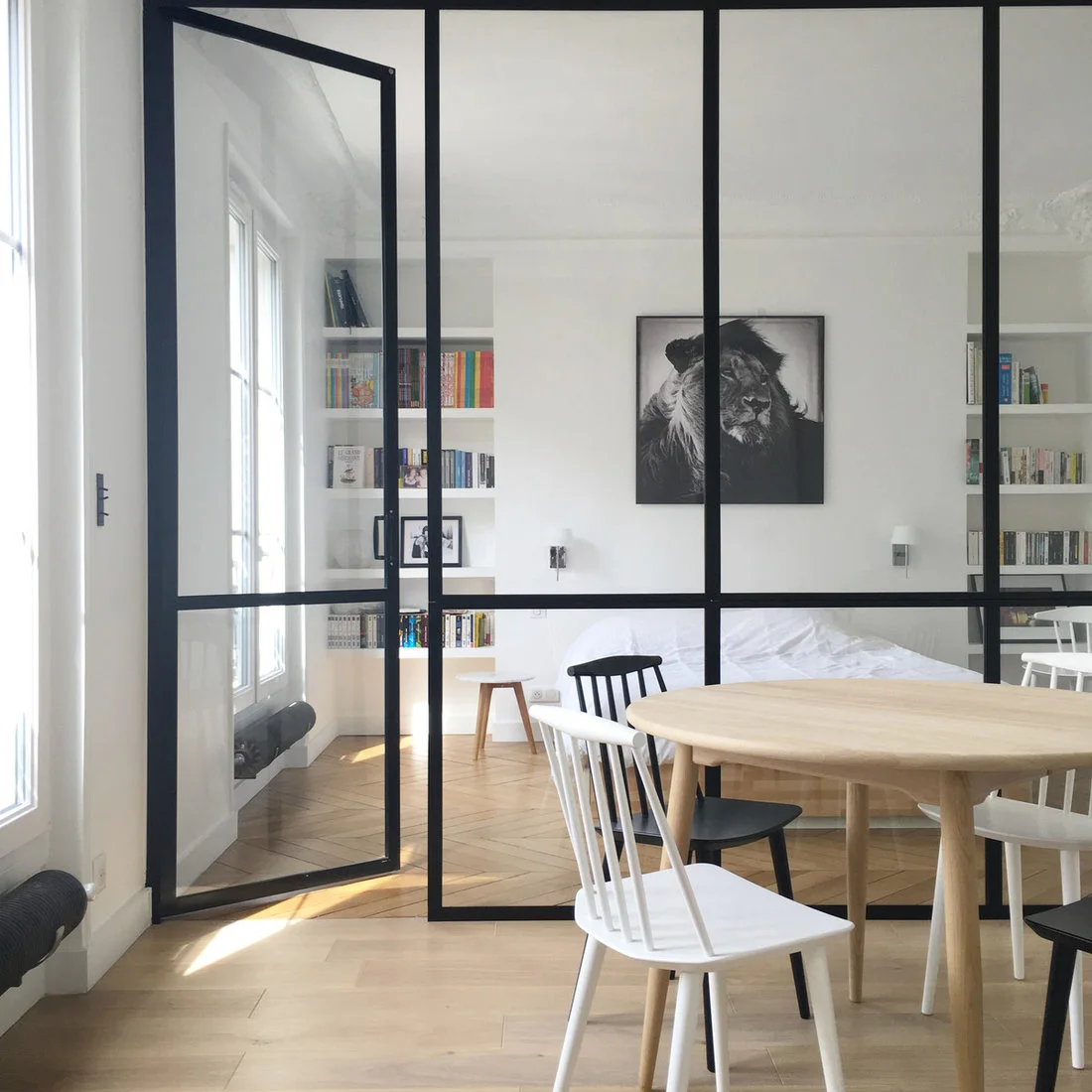 A pale oval wood table and black-and-white spindle chairs stand in front of a black-framed glass bedroom partition.