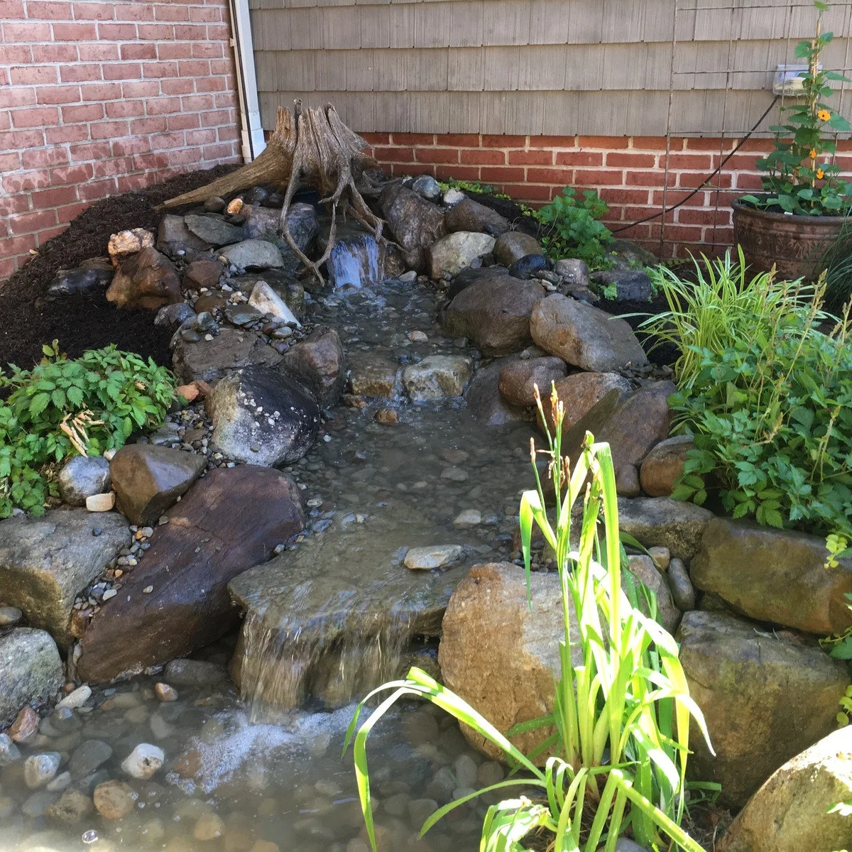 A short rock cascade in a corner beside brick walls, with a weathered stump at the top and a shallow pebble-lined run below.