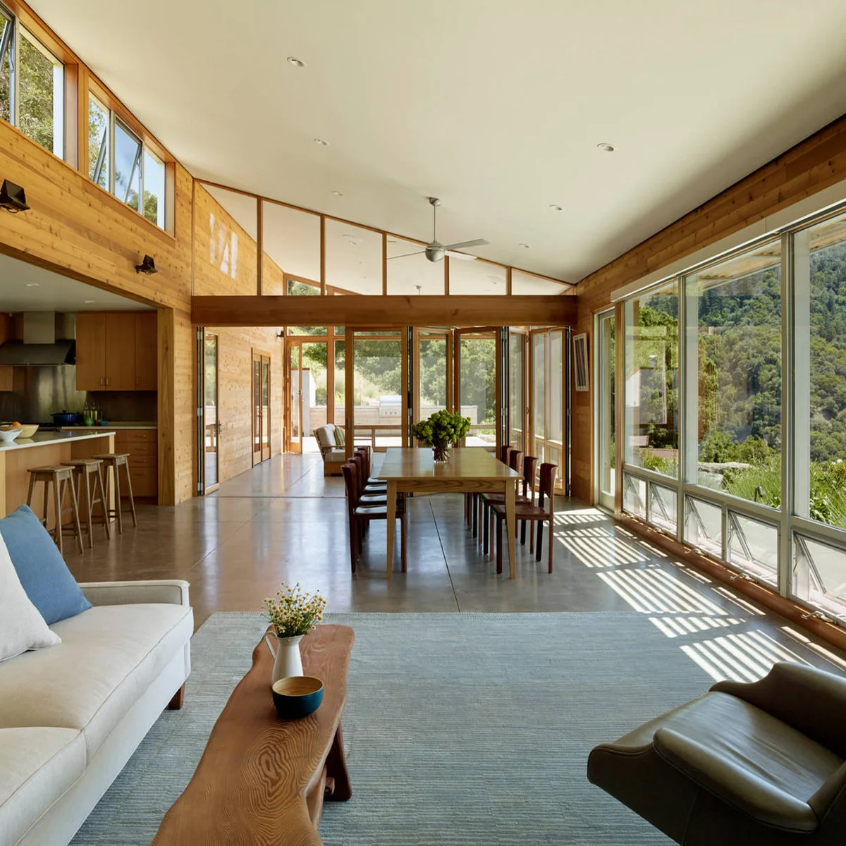 Open living room with a pale sofa, long wood coffee table, dining table and kitchen beside tall windows