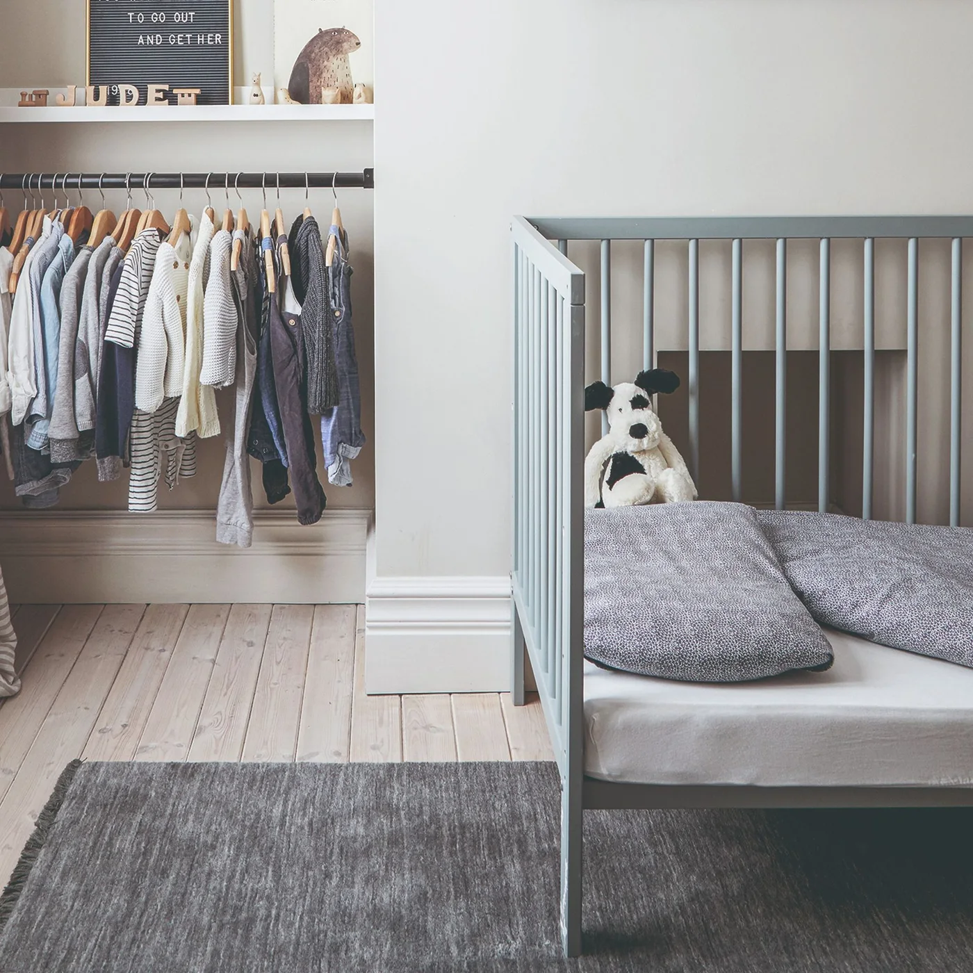 A small gray child’s bed stands beside an open clothes rail below a white shelf, with pale walls, a gray rug and visible wood flooring