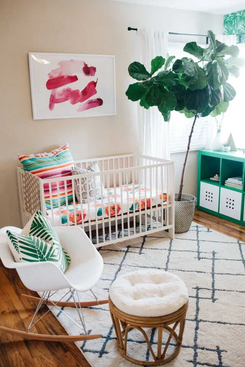 Pale nursery with white crib, brightly striped staged cushion, green storage, leafy plant, white rocking chair and geometric rug.