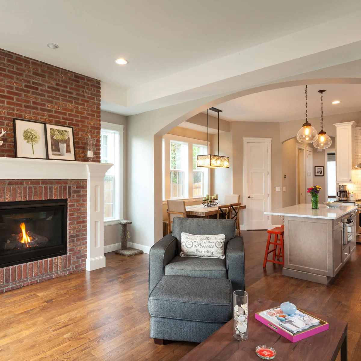 A red brick fireplace has a pale paneled mantel beside gray walls, wood flooring and a gray chair in an open living space.