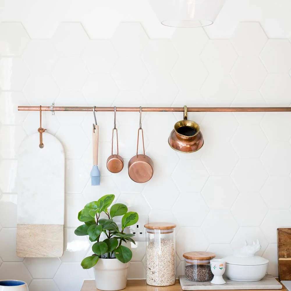 White hexagonal tiles form a kitchen wall behind a copper-toned utensil rail, pale canisters and a small plant