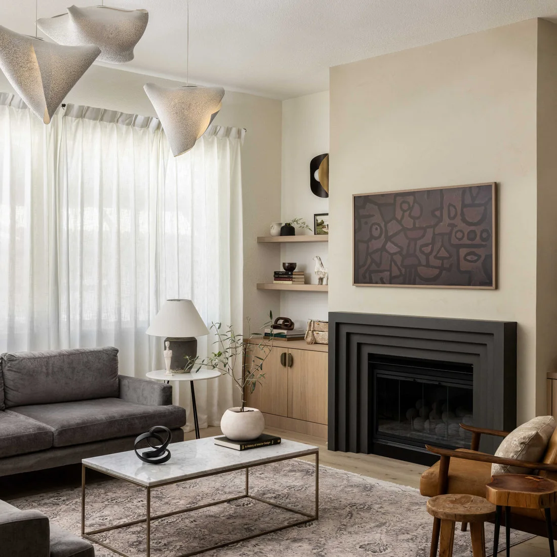 A black stepped fireplace surround sits beneath muted abstract artwork in a cream room, beside wood built-ins, gray seating and a small metal-framed coffee table.