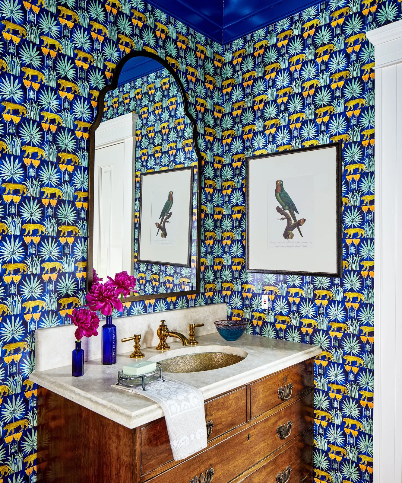 Blue wallpaper with yellow animal and pale leaf motifs surrounds a wood vanity, shaped dark mirror, framed bird picture and warm-metal basin fittings.