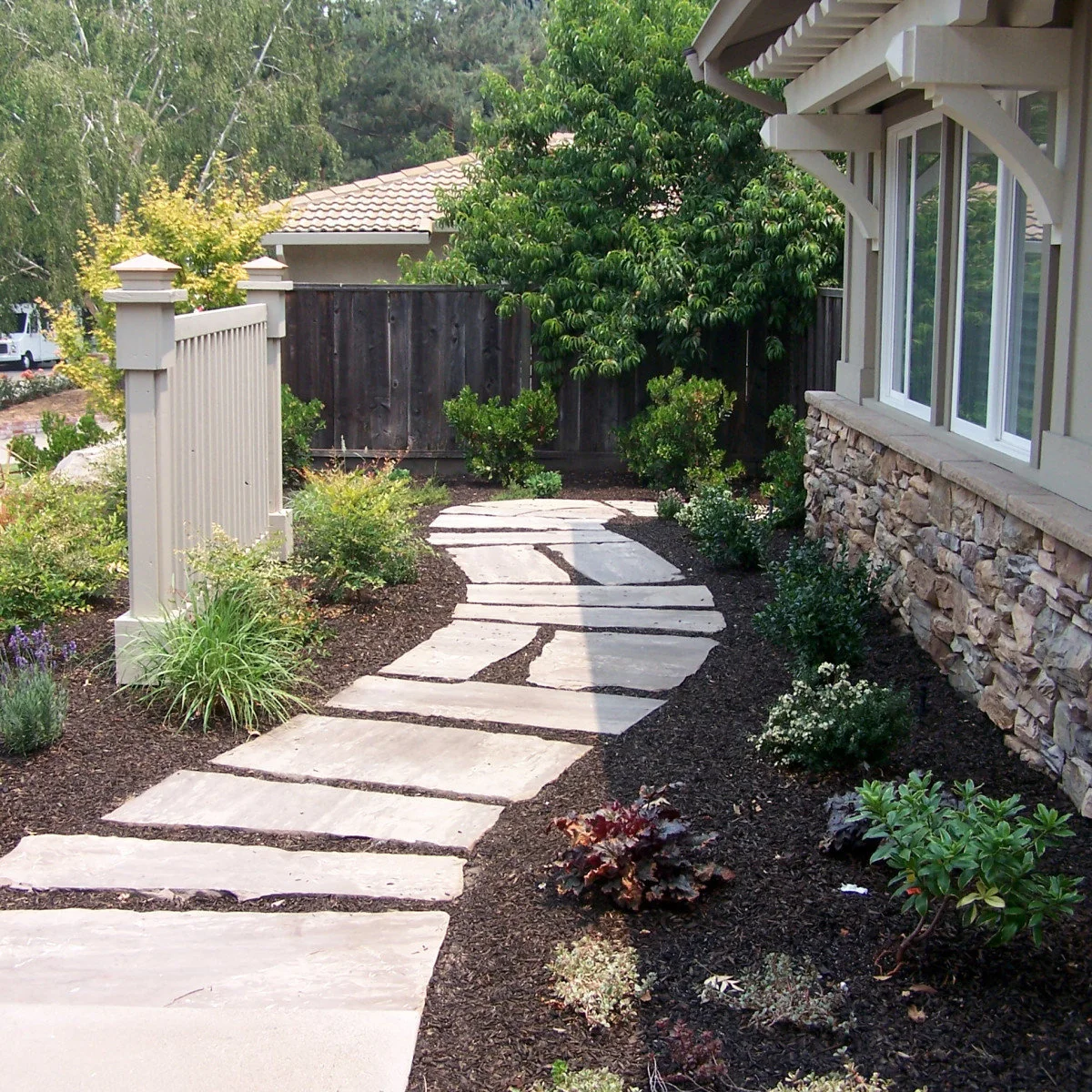 A curving path of broad pale slabs through mulched planting beside a house and a short white fence.