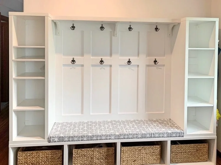 White mudroom arrangement with tall open side units, two rows of black hooks, a patterned cushion and lower woven baskets.