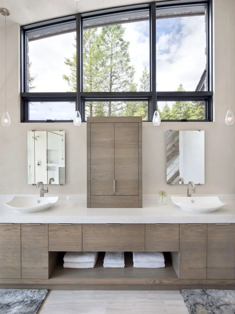 Two white vessel basins sit on a long pale counter with wood-fronted cabinets, a central raised cupboard, open towel storage and high black-framed windows