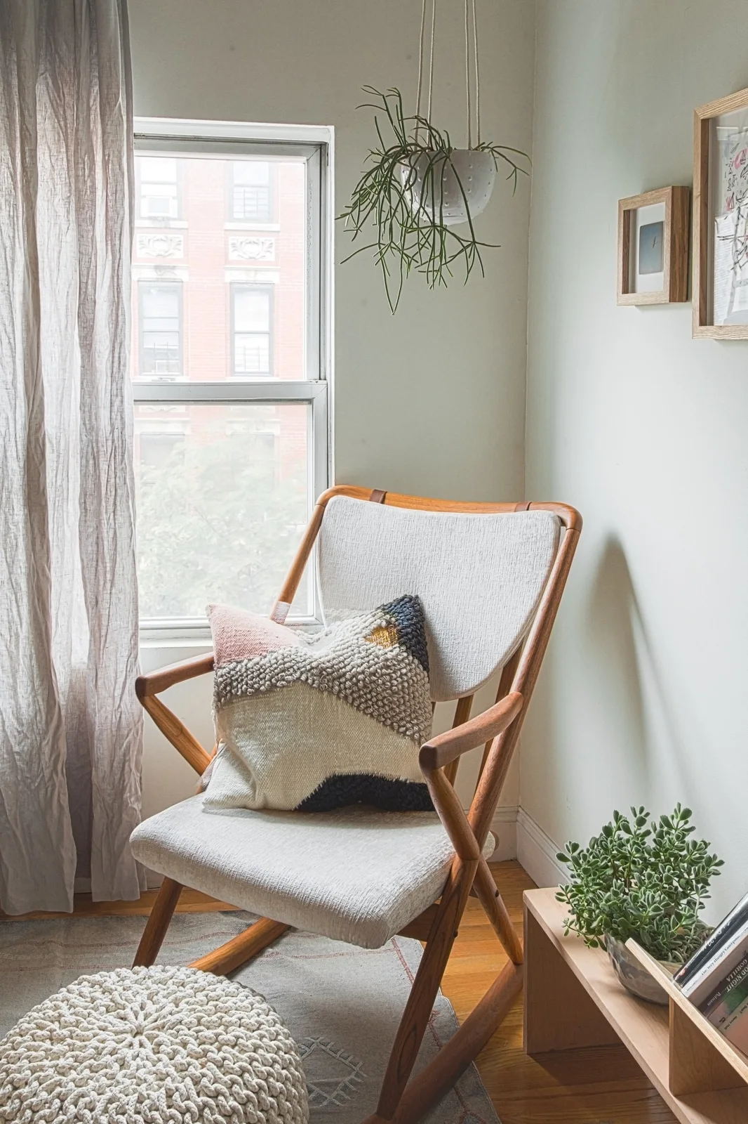 A timber-framed nursery rocking chair sits near a window beside a low shelf and a knitted round footrest.