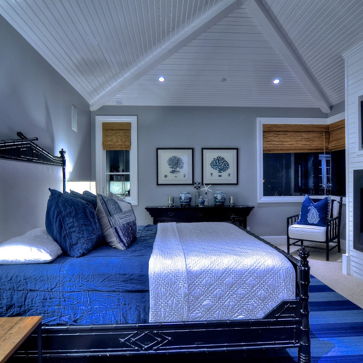 Blue quilt and pillows on a dark framed bed beneath a white vaulted ceiling with woven window shades.